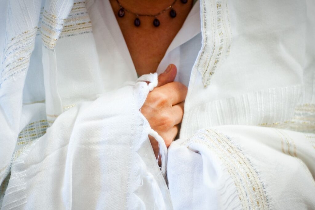 A person drapes a white, embroidered tallit over their shoulders, hand resting on their chest.