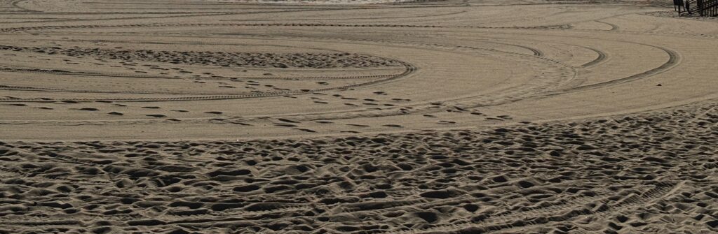 Curved tire tracks and footprints are visible in the sand on a sunny beach.