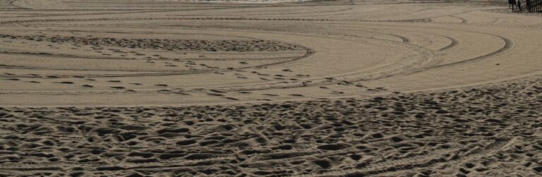 Curved tire tracks and footprints are visible in the sand on a sunny beach.