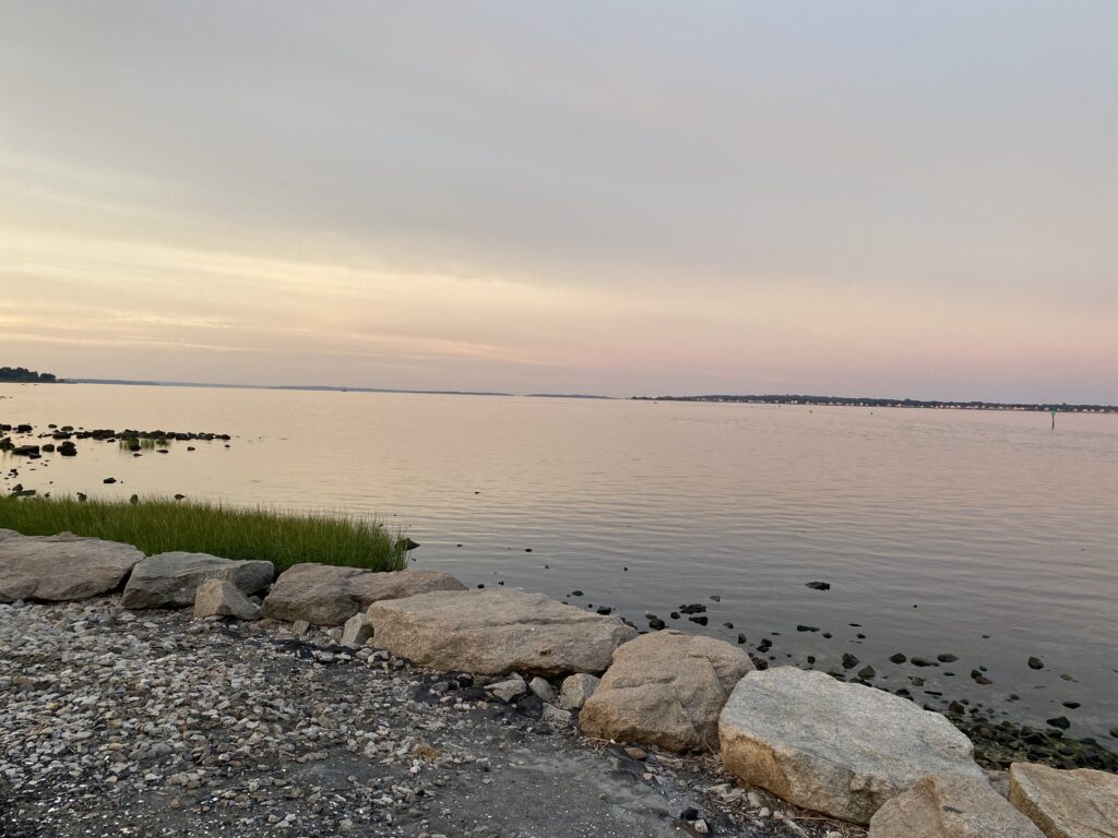 Rocky shoreline with calm water at sunset, sky glowing with soft pink and purple hues.
