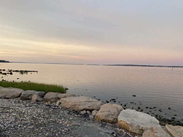 Rocky shoreline with calm water at sunset, sky glowing with soft pink and purple hues.
