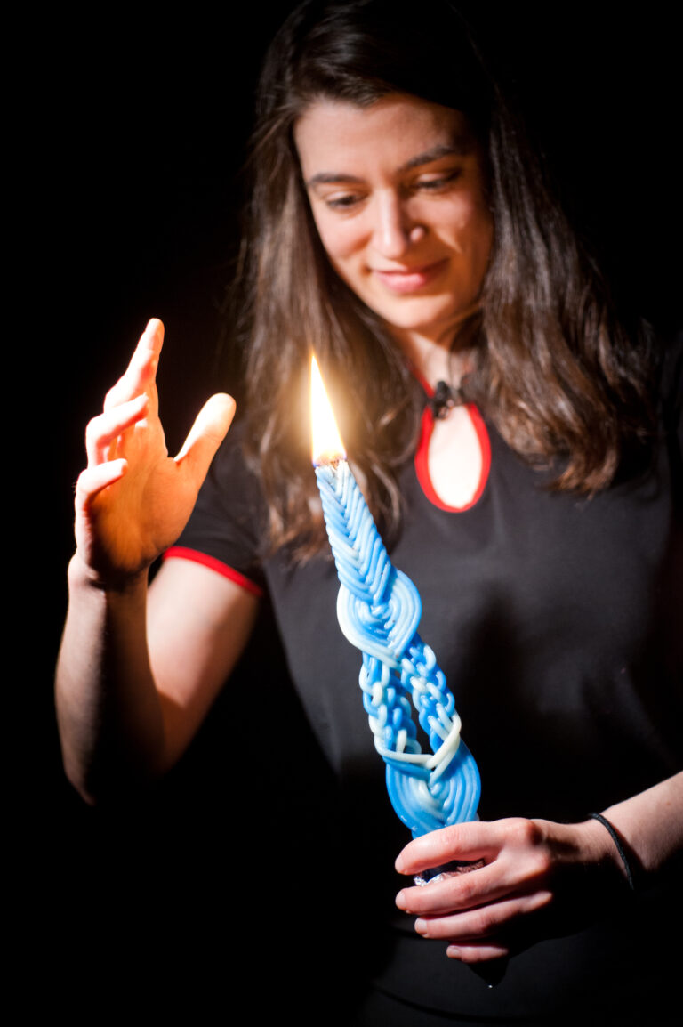 A woman holds a lit blue braided candle, raising her hand toward its flame against a dark background.
