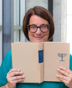 Dr. Joellyn Zollman has short brown hair, glasses and is holding a book