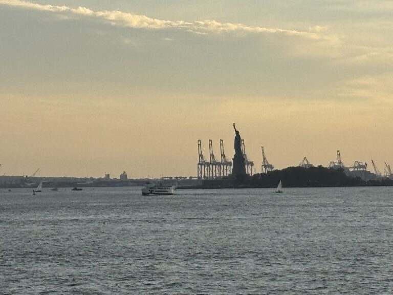 The Statue of Liberty and cranes are seen at sunset across the water, with boats and a cloudy sky.