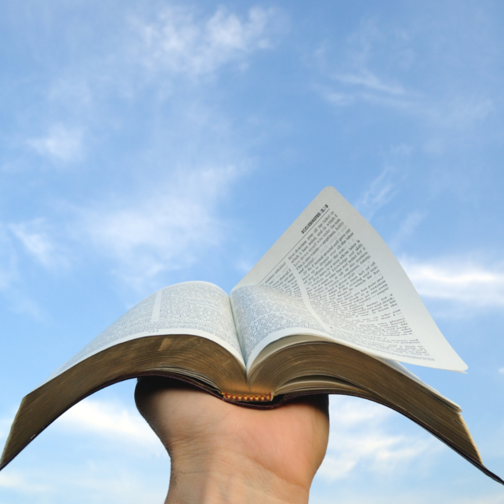 A hand holds an open book up to a blue sky with scattered clouds.
