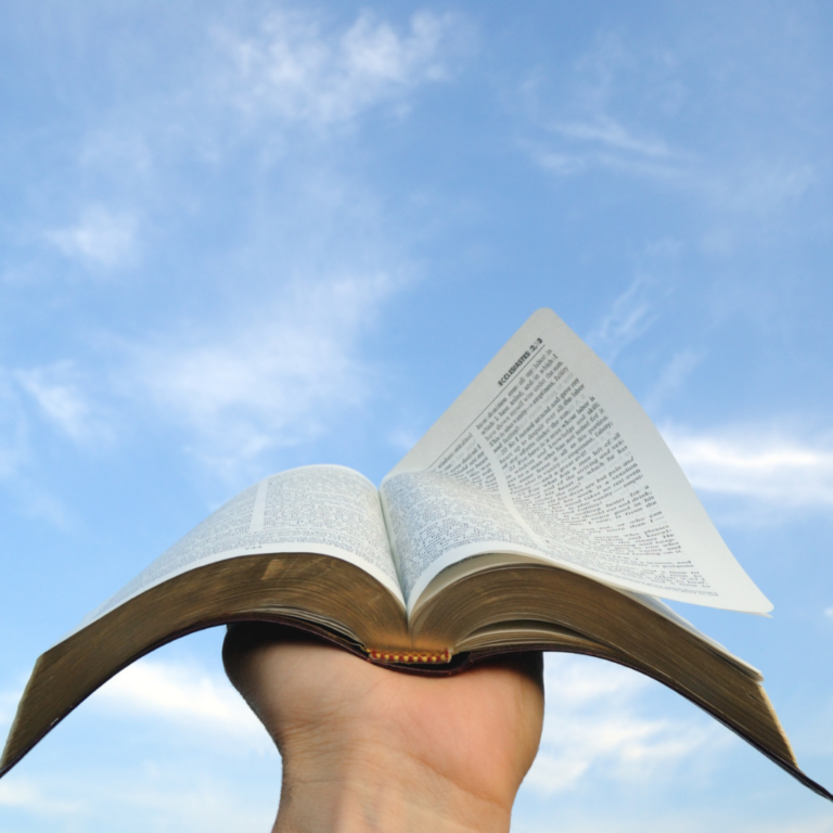 A hand holds an open book up to a blue sky with scattered clouds.