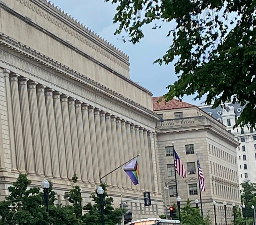 A building with columns, U.S. flags, and a Pride flag flying, with trees in the foreground.