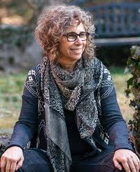 Rabbi Yael Levy is a smiling woman with curly hair and glasses sits outdoors, wearing a patterned scarf and dark sweater.
