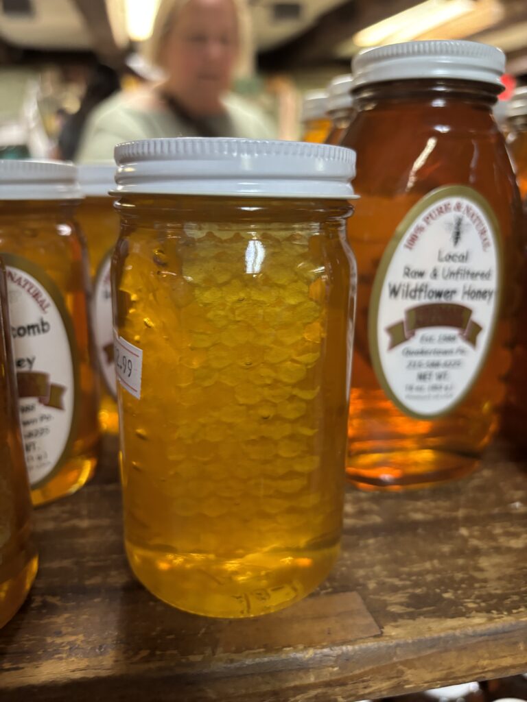 A jar of honey with honeycomb inside, displayed on a wooden shelf among other honey jars.