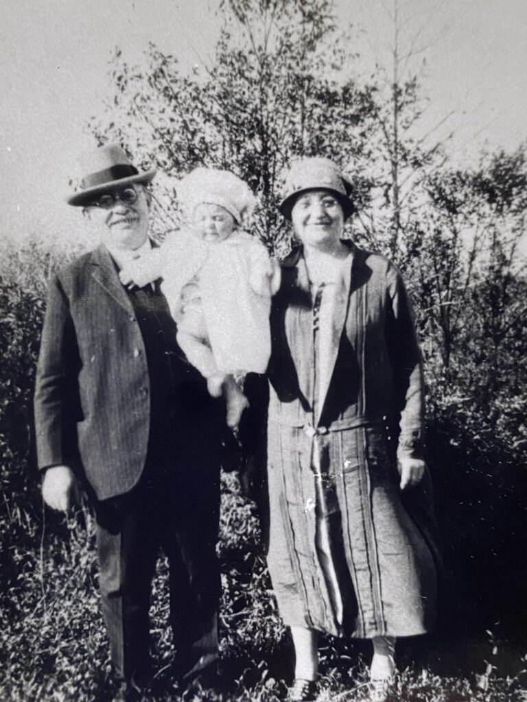 Black and white photo of two adults and a baby standing outdoors, all dressed in vintage clothing.