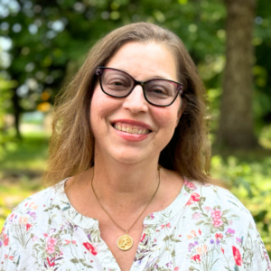 Gabrielle is a smiling woman with glasses and floral shirt, standing outdoors with trees and greenery in the background.
