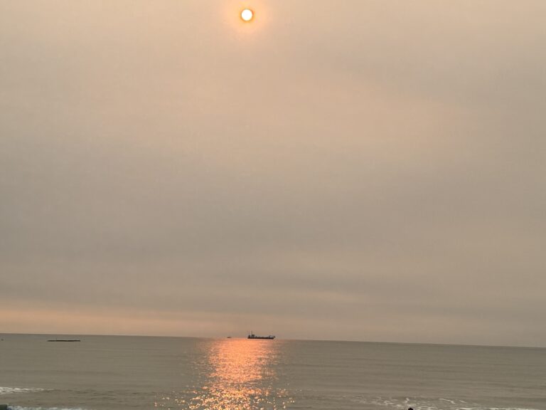 The sun glows orange over a calm ocean with a distant ship and shimmering reflection on the water.
