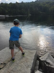 A boy in a blue shirt and camo shorts fishes from rocks by a lake surrounded by trees.