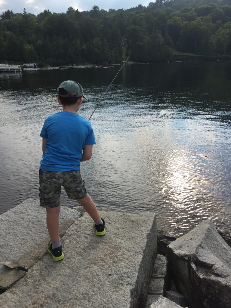 A boy in a blue shirt and camo shorts fishes from rocks by a lake surrounded by trees.