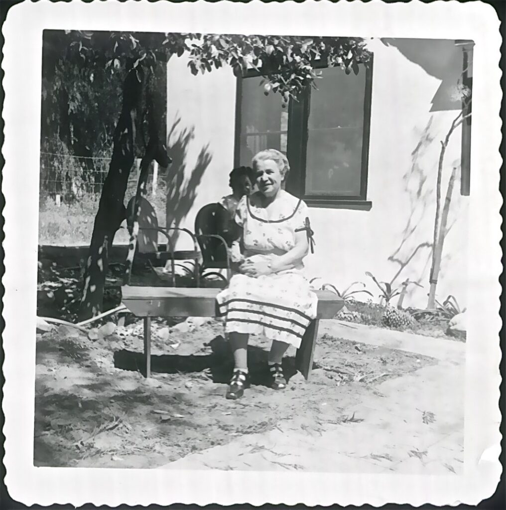 Elderly woman in a dress sits on a bench outside, with a young child behind her near a house.