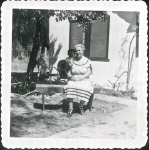 Elderly woman in a dress sits on a bench outside, with a young child behind her near a house.