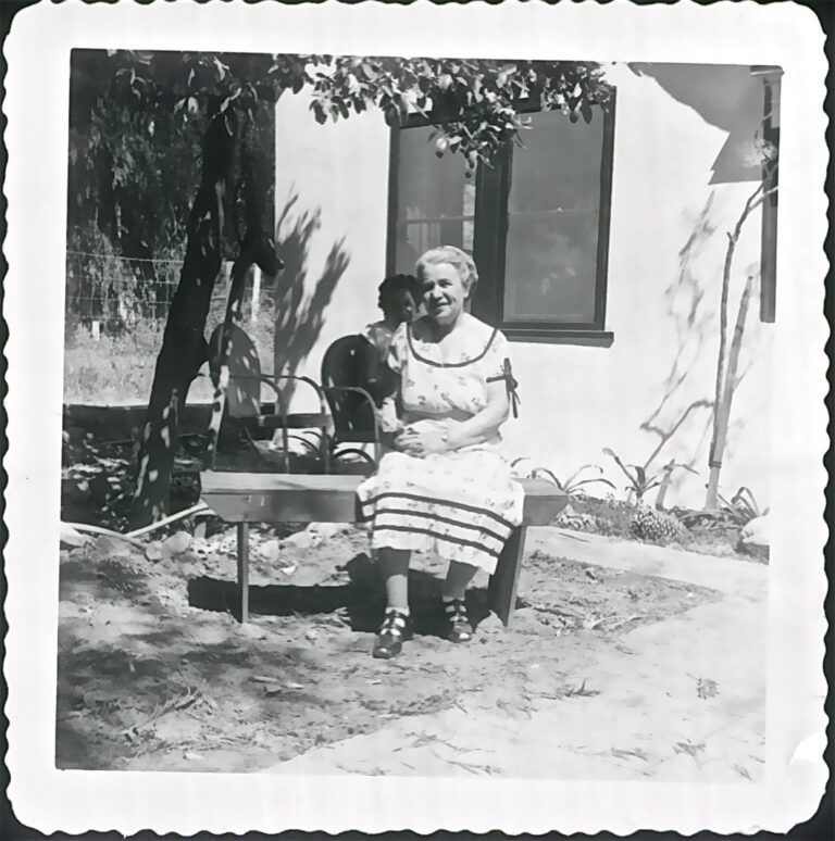 Elderly woman in a dress sits on a bench outside, with a young child behind her near a house.