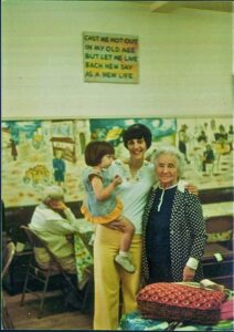 Three women, one holding a child, smile in a colorful room with a sign and mural in the background.