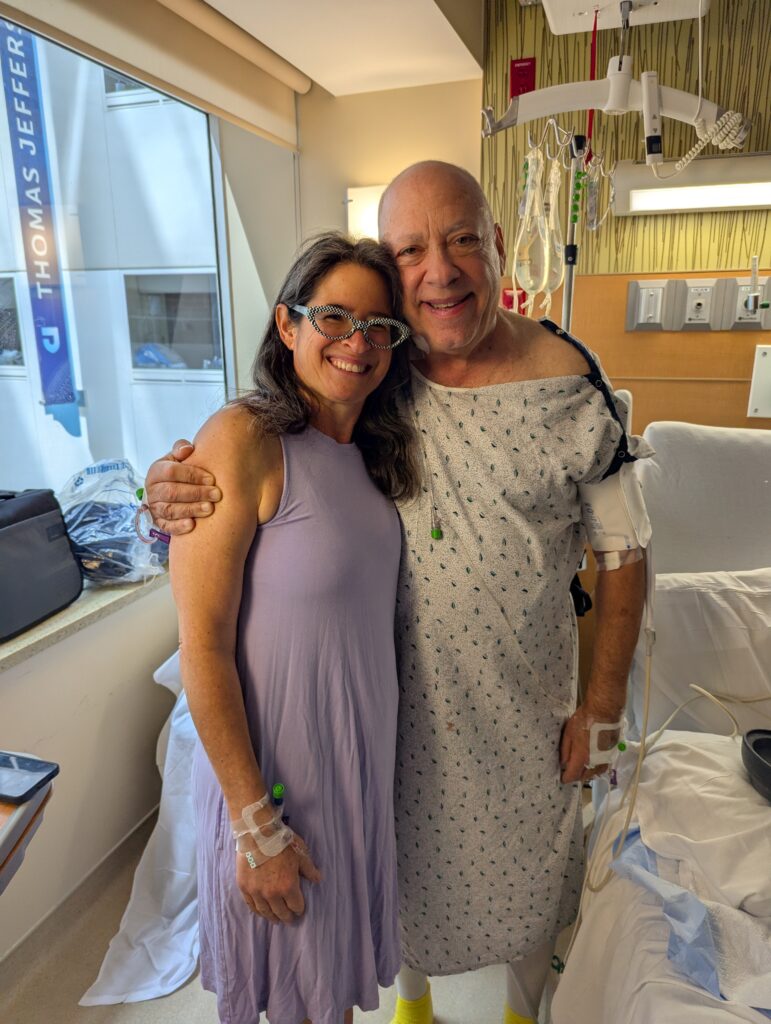 Smiling woman hugs man in hospital gown with medical equipment, both standing in a bright hospital room.