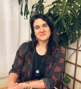 Woman with long dark hair sits smiling, wearing a patterned shirt, blue pendant, and gold bracelets. Plants behind.