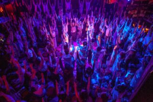 A crowd of people under colorful lights raise their arms together at a lively indoor event or concert.