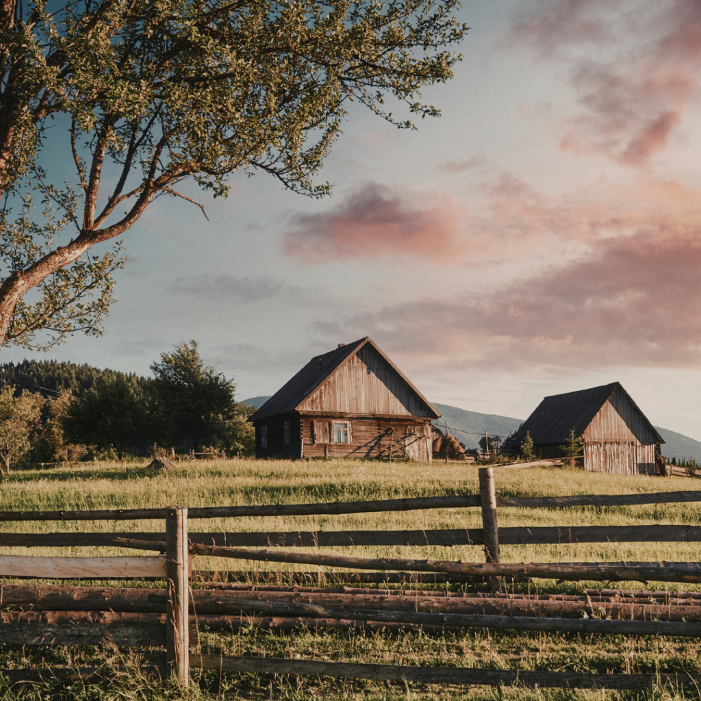 Two wooden houses sit in a grassy field with a fence and a tree under a cloudy sky at sunset.