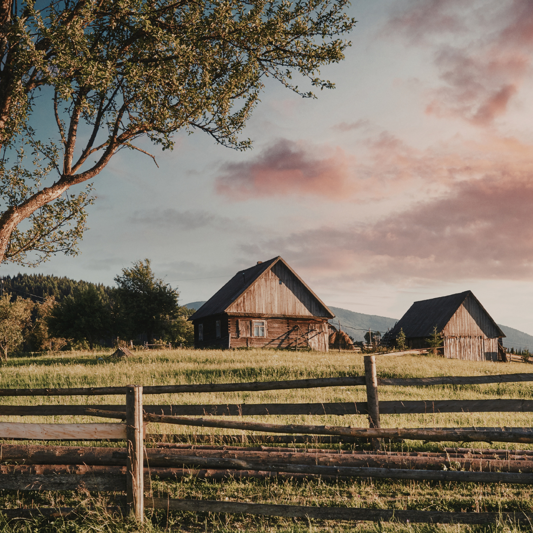 Two wooden houses sit in a grassy field with a fence and a tree under a cloudy sky at sunset.