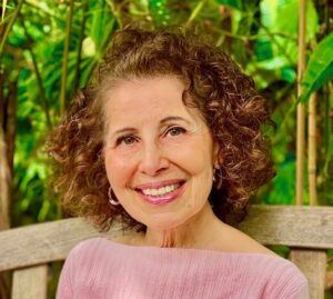 Smiling woman with curly hair sits on a wooden bench in a lush, green outdoor setting.