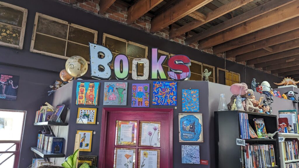 Colorful Books sign above bookshelves, surrounded by art, books, and unique objects in a cozy bookstore.