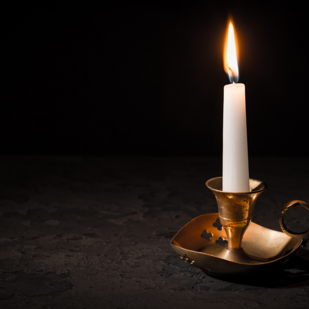 A lit white candle in a brass holder sits on a dark textured surface with a black background.