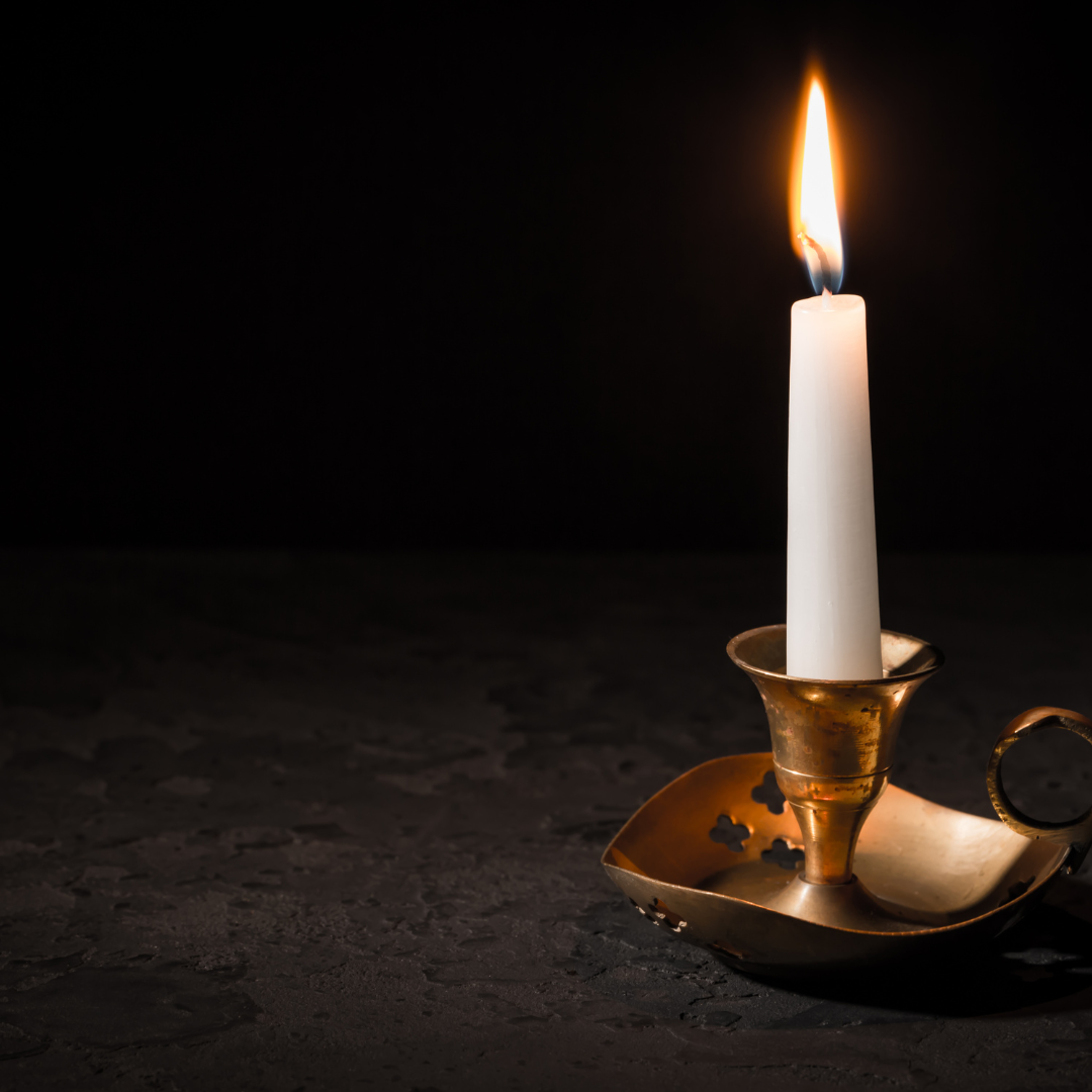 A lit white candle in a brass holder sits on a dark textured surface with a black background.