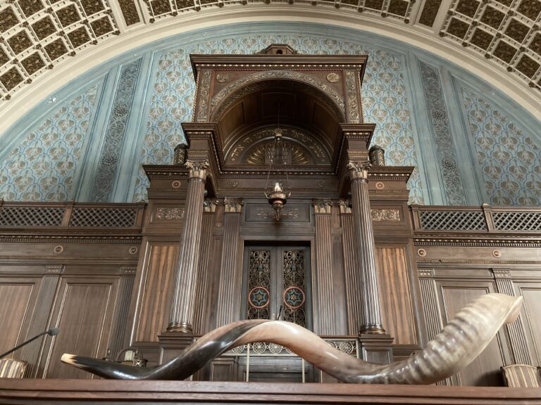 A curved shofar rests on a wooden surface in front of an ornate synagogue ark and blue mural.