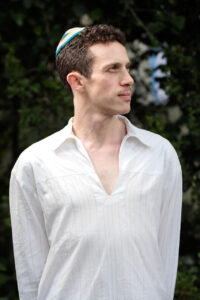 A man in a white shirt and colorful kippah stands outdoors, looking to the side.