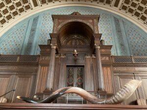 Curved shofar on a wooden bimah in front of an ornate ark in a synagogue with blue patterned walls.