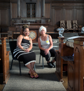 Two women sit side by side in a wood-paneled room with ornate details and large chairs in the background.