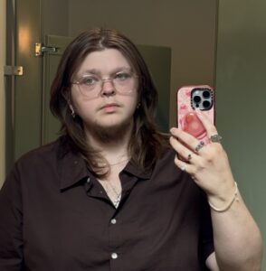 Person with long hair and glasses takes a mirror selfie in a bathroom, wearing a brown shirt.