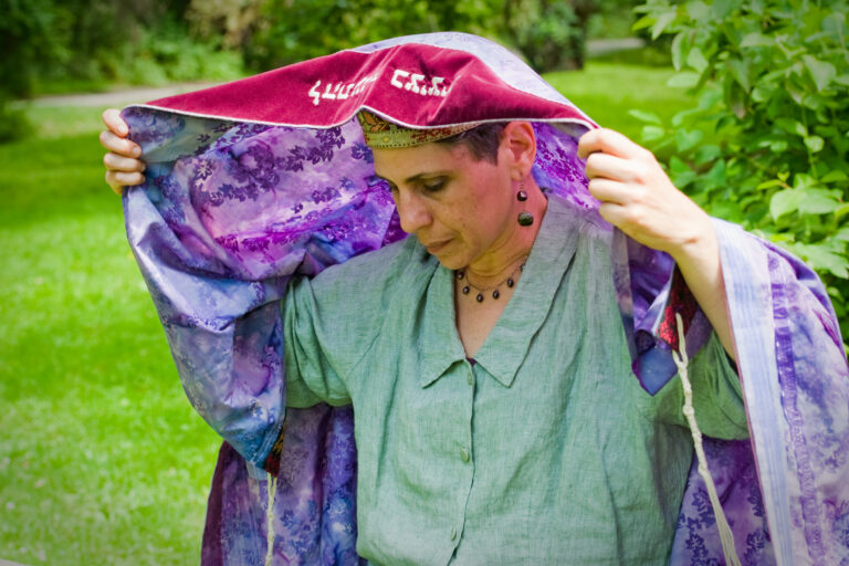 Person putting on a purple and red prayer shawl outdoors, with green grass and trees in the background.