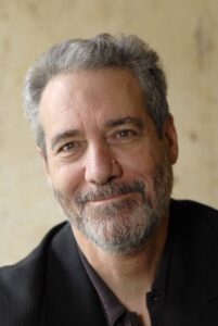 Smiling middle-aged man with gray hair and beard wearing a dark jacket, posed against a light background.