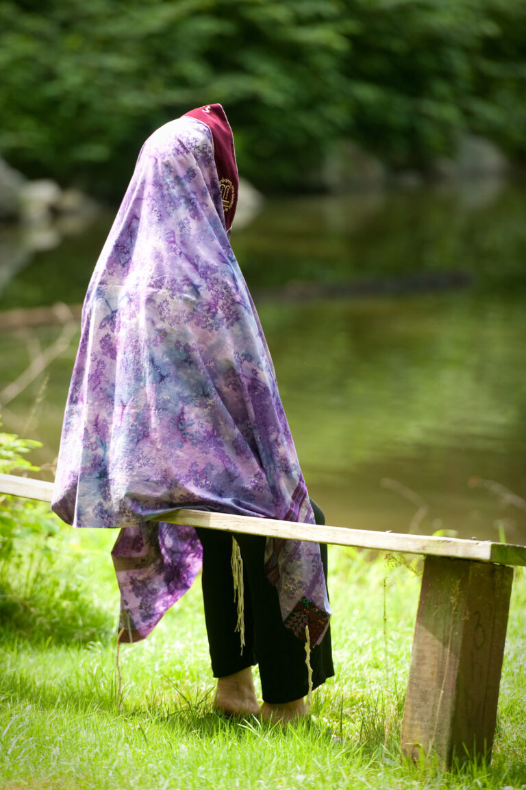 A person in a purple floral shawl stands on grass by a wooden bench near a pond.