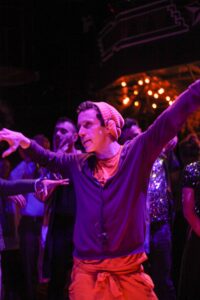 Man in orange beanie and cardigan dances energetically in a lively, dimly lit crowd.