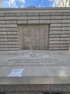 A stone memorial with engraved Hebrew text stands in front of a gray building under a partly cloudy sky.