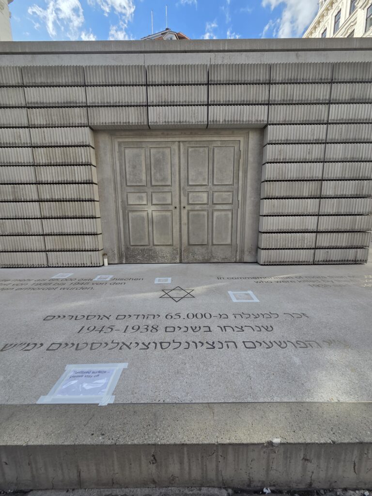 A stone memorial with engraved Hebrew text stands in front of a gray building under a partly cloudy sky.