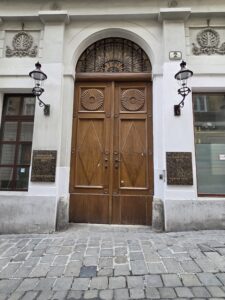 Tall wooden double doors with carvings, flanked by lamps, on a cobblestone street.