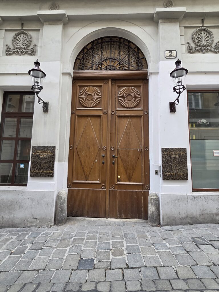 Tall wooden double doors with carvings, flanked by lamps, on a cobblestone street.