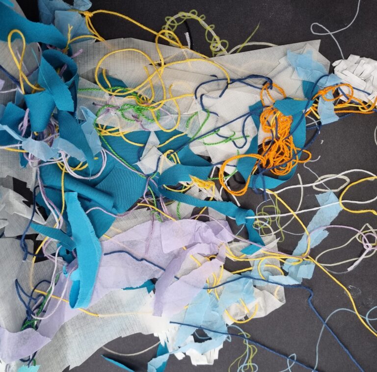 Colorful fabric scraps and threads are scattered on a dark surface, creating an abstract, tangled composition.