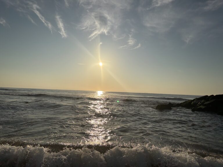 Sunset over the ocean, waves gently crashing near rocks, and clouds scattered across the sky.