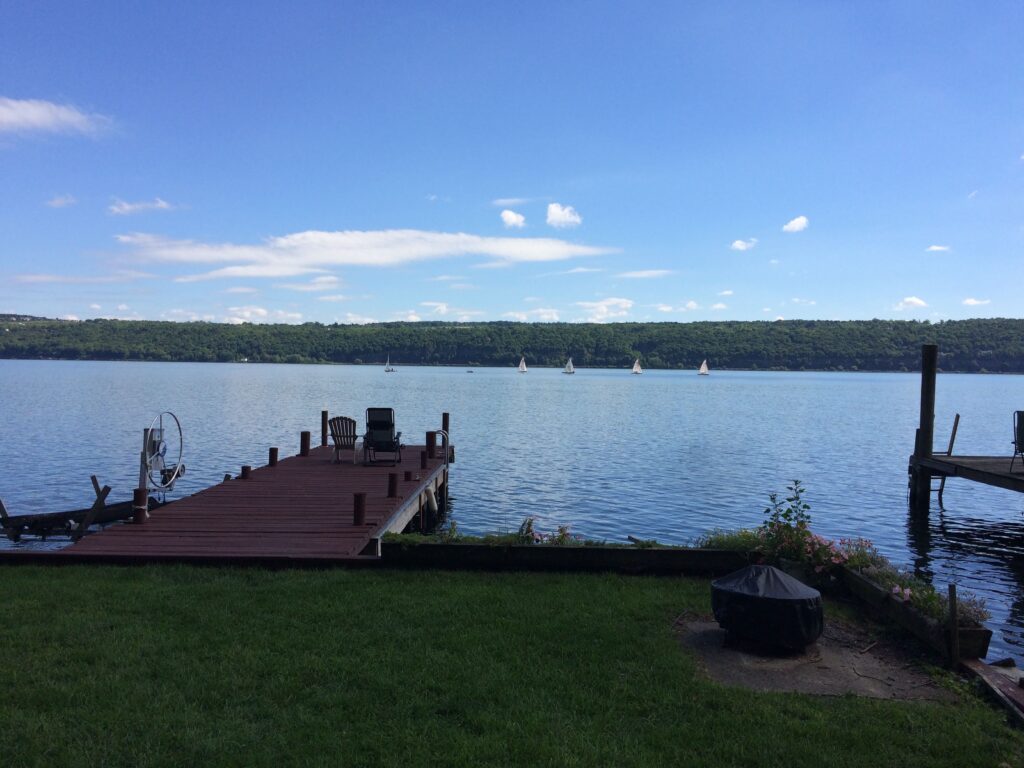 A wooden dock extends into a calm lake with sailboats in the distance and green hills beyond.