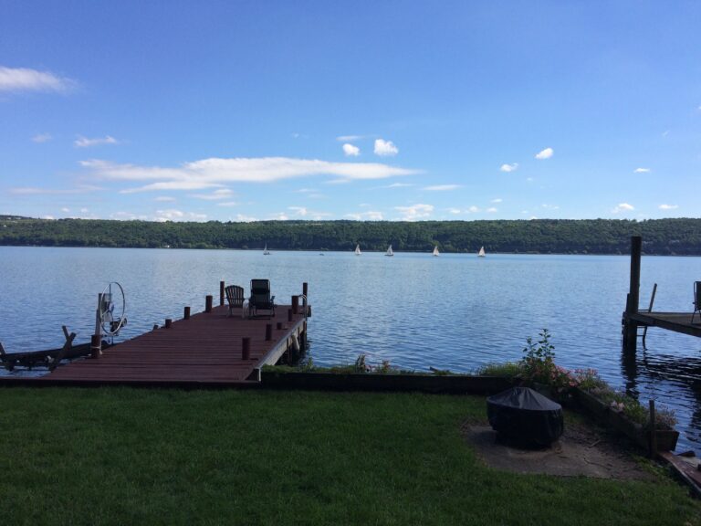 A wooden dock extends into a calm lake with sailboats in the distance and green hills beyond.