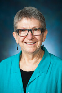 Smiling older woman with short gray hair, glasses, teal shirt, and earrings against a blue background.