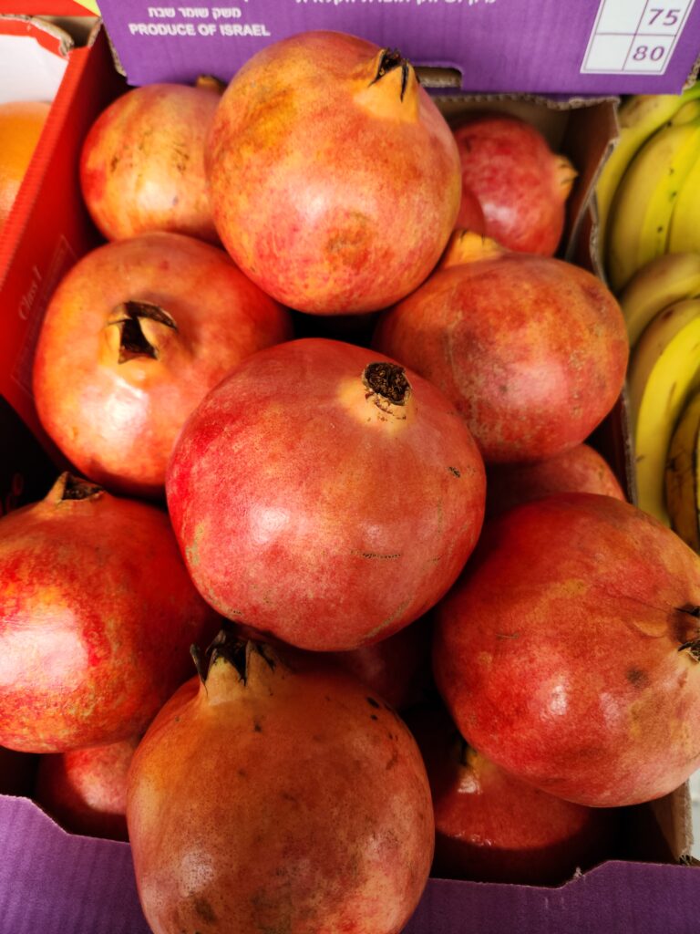 A pile of ripe pomegranates fills a box, with yellow bananas visible in the background.